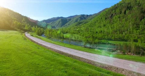 UHD 4K aerial view. Low flight over fresh cold mountain road, river and meadow at sunny summer morning. Green trees and sun rays on horisont.