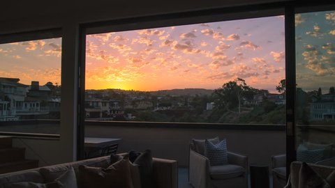 Early morning time lapse from a luxury home living room through balcony doors of a stunning yellow and gold sunrise with clouds overhead overlooking a canyon, coastal homes and mountains.