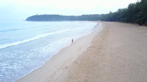 Young adult man rest after running, walk along empty tropical beach, early morning time. Aerial camera follow behind, wide angle shot. Fresh air from sea, clear empty sand area, healthy environment