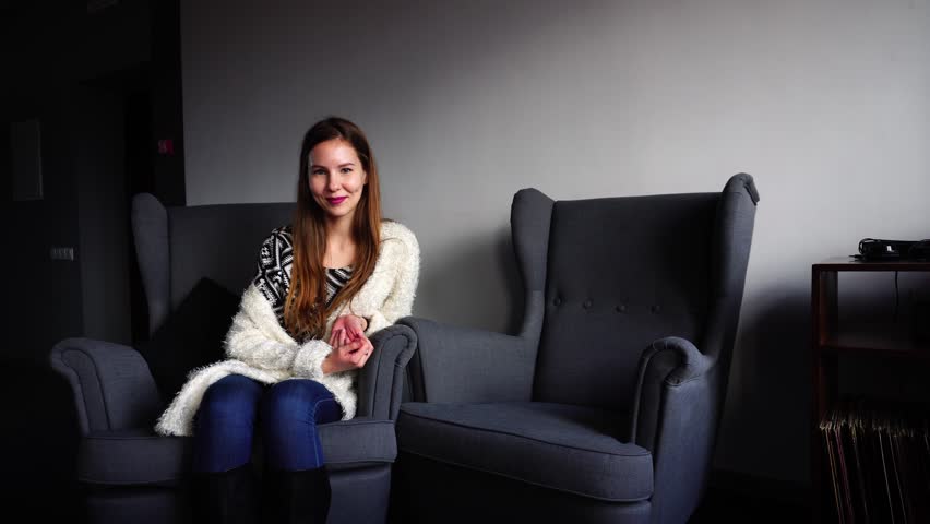 Cute young woman smiles and looks at camera, head over arm, posing, sitting on gray armchair in stylish cafe and wearing printed t-shirt, fluffy white cardigan and blue jeans with high black boots