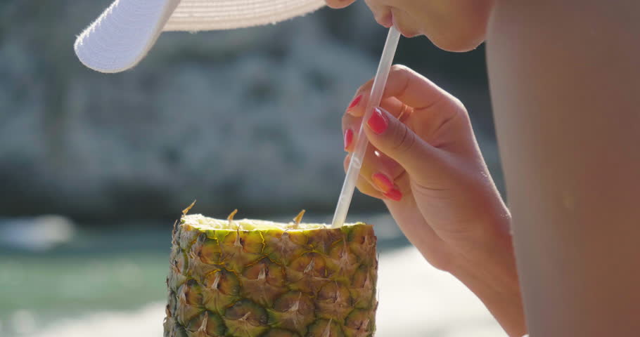 Beautiful young girl drinks a pineapple cocktail at the sea, in a swimsuit in a white hat, golden sand, sea background. Concept: sea rest, clean air, freedom, sun, travel, waves, fresh fruit, vacation