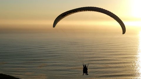 Paragliding into sunset seascape, with pilot adjusting wing controls, changing direction, sunset lit ocean waves, sun beam trail to horizon, two small distant boats, yellow orange light, horizon