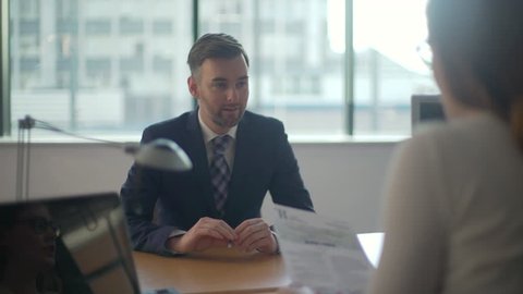 Man goes to job interview, sits and desk and is being interviewed by female boss, nervous and anxious he fiddles with a pen as she checks over his CV.