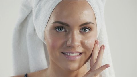 Portrait of happy smiling beautiful young woman touching skin or applying cream, after shower with towel on head