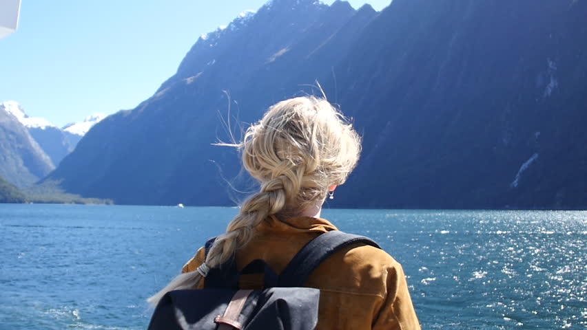 Woman Gazes at Mountain Lake - A blonde woman shown from behind looks out over a lake in front of a mountain, and then turns her head to the side; the camera then pans to the right to show mountains Royalty-Free Stock Footage #29563858