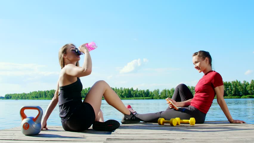 Two beautiful, athletic blonde girls, women have rest after training outdoors, drink water, talk, laugh. Lake, river, blue sky and forest in the background, summer sunny day