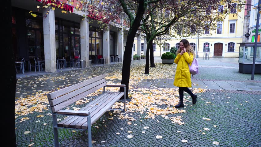 Fine girl talks on phone and goes to wooden bench, sits down and reports  affairs and impressions about city and excursion to  friend. Young European-looking woman with long blond hair smiles and si
