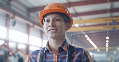 Tilt down of beautiful female factory worker walking through industrial building, holding welding mask and smiling