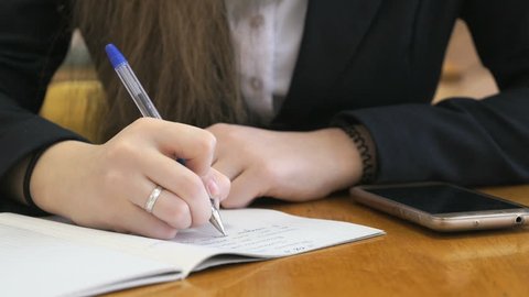 Student dressed in a black suit sits at a school desk writing text in exercise book using ballpoint pen next to a golden mobile phone. Close-up
