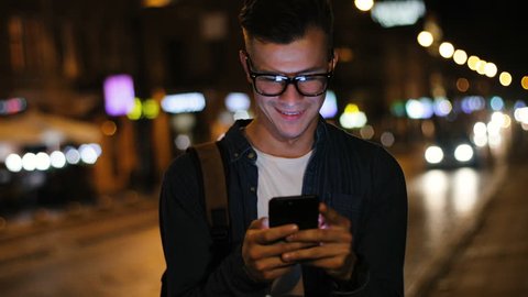 Close up shot of attractive man in the stylish glasses using smart phone in the street at the evening time on the light background.