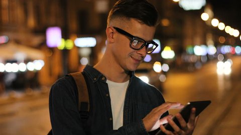 View from the side of emotional man in the stylish glasses using smart phone in the street at the evening time on road background.