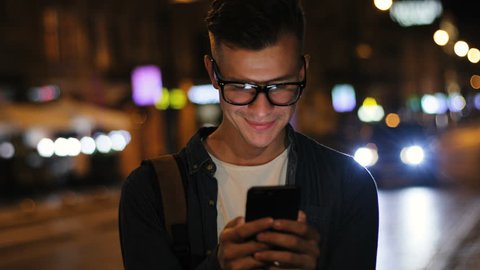 Emotional man in the stylish glasses using smart phone in the street at the evening time on road background.