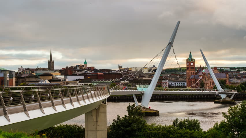 Derry, Ireland. Illuminated Peace bridge in Derry Londonderry in Northern Ireland with city center at the background. Time-lapse from day to night with cloudy sky, reflection in the river.