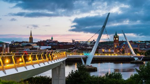 Derry, Ireland. Illuminated Peace bridge in Derry Londonderry in Northern Ireland with city center at the background. Time-lapse from day to night with cloudy sky, reflection in the river.