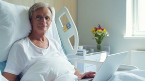 Positive aged man lying in the hospital bed with laptop