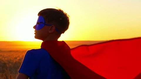 Kid boy dressed in a superhero costume staring into the distance at sunset.