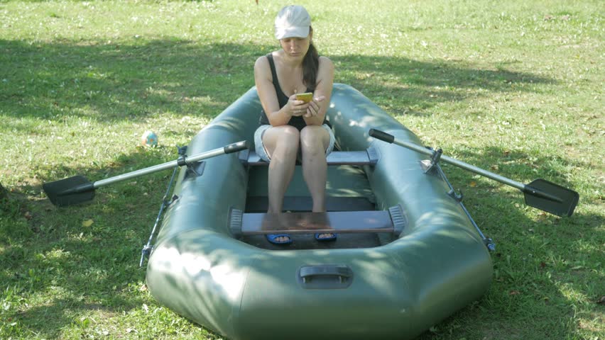 A pretty woman sits in an inflatable boat near the house and dials the phone. Shorts and a white baseball cap on the head