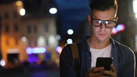 Attractive man in the stylish glasses using smart phone in the street at the evening time on the light background of the bulding.
