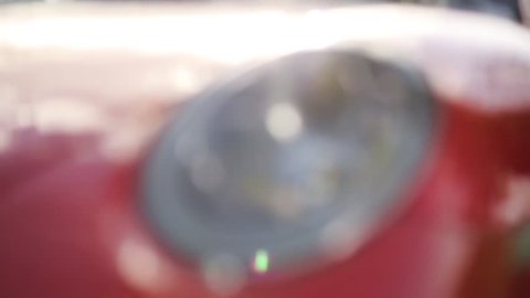 The headlight of an expensive red sports car, the sun reflects on the car's body, it looks expensive, the car stands on a country road
