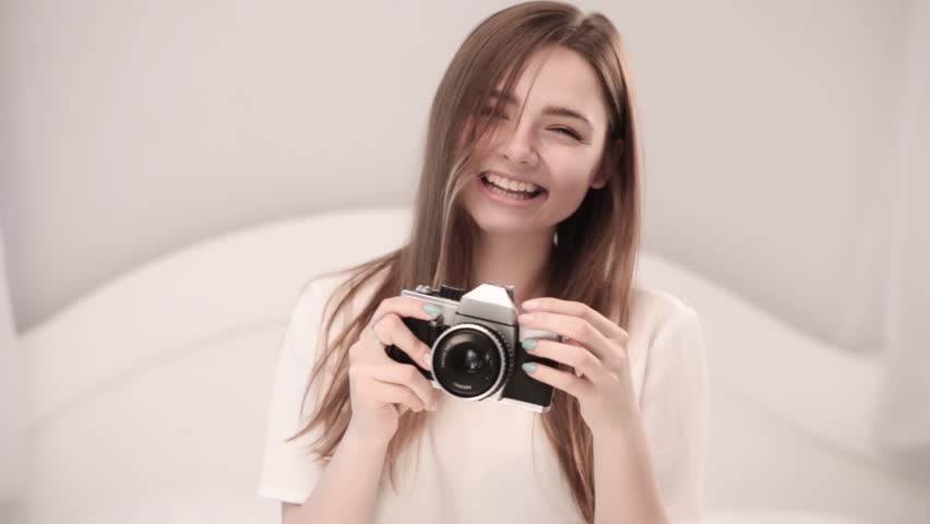 Close-up of the girl, she sits on a light bed and holds an old camera in her hand, the girl is inspired to take pictures in front of her, emotions are playing on her face
