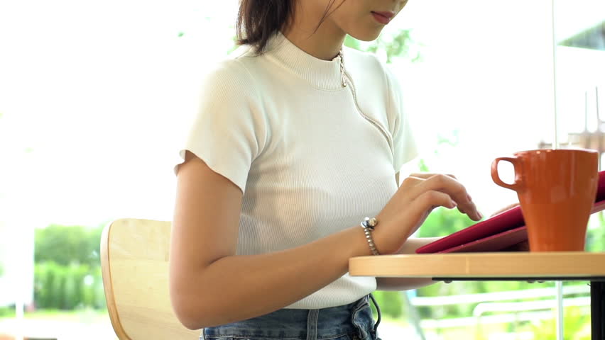 Young Asian Teenager having a relaxing time in a coffee shop typing and chatting on a computer tablet on a bright sunny day