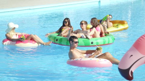 SLOW MOTION LENS FLARE: Playful friends enjoying on colorful floaties in pool. Cheerful young people drinking and having water gun fight on inflatable pizza, flamingo, watermelon and doughnut floats