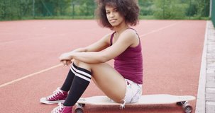 Side view of fit woman in shorts sitting on longboard on sports ground for basketball and looking confidently at camera.