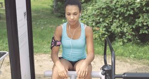 Charming ethnic girl in sportswear listening to music with smartphone while training abdominal muscles in park gym.