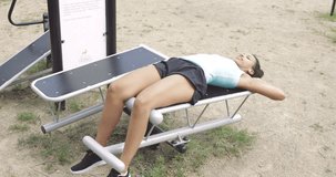 Fit ethnic girl in sportive shorts listening to music while working out and training abdominal muscles on bench in park and looking forward.
