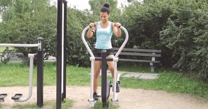 Confident young woman in sportive clothing working out on exerciser in street gym while listening to music with smartphone and looking away.