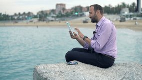 Young, happy businessman chatting on tablet sitting on rock near sea 
