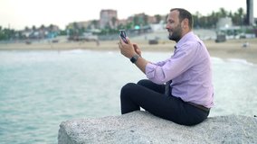 Young, happy businessman chatting on smartphone sitting on rock near sea 
