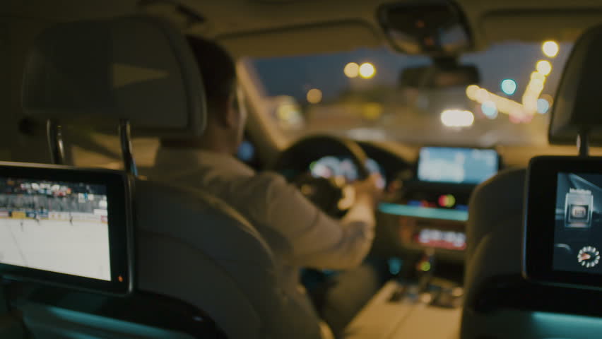 Man driving at night in the city a modern car, TV showing a Ice hockey game on the back of his car seat.