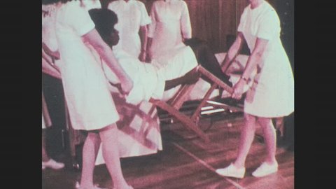 1970s: Two women nurses carry a woman tied to chair and set her down. Man in uniform talks to medical staff. Barefoot person in medical gown limps across linoleum floor.