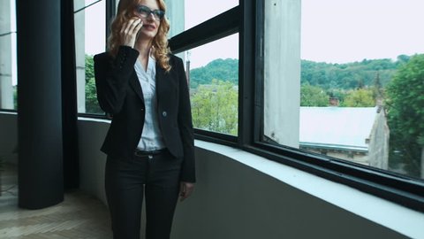 Beautiful adult businesswoman talking on phone,smiling, walking in office near big window. Business, communication concept