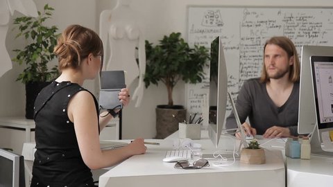 In office woman shows a man a notebook with notes at her desk. Two employees at a white table with computers solve problem according to plan on paper of diary.