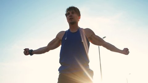 Young handsome man athlete in blue t-shirt and headphones doing warm up before running at sunset