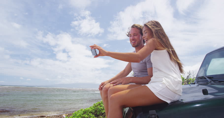 Happy couple taking selfie using smartphone at Shipwreck beach, Lanai. Young tourists are sitting on offroad vehicle trunk enjoying summer vacation on Lanai, Hawaii, taking phone pictures.