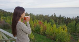 Young woman looking at trees and sea while standing on balcony in forest chalet or cabin. Cozy woman drinking coffee while enjoying view of fall forest on vacation during autumn.