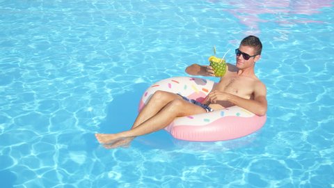 SLOW MOTION CLOSE UP: Happy guy sipping pineapple drink and relaxing on inflatable doughnut float. Young man enjoying summer vacation drinking cocktails on fun doughnut floatie in pool