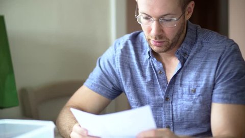 A man in debt is worried and stressed whilst looking through bill letters. Sad and anxious he holds his head in hands. White Caucasian wearing glasses in 30's.