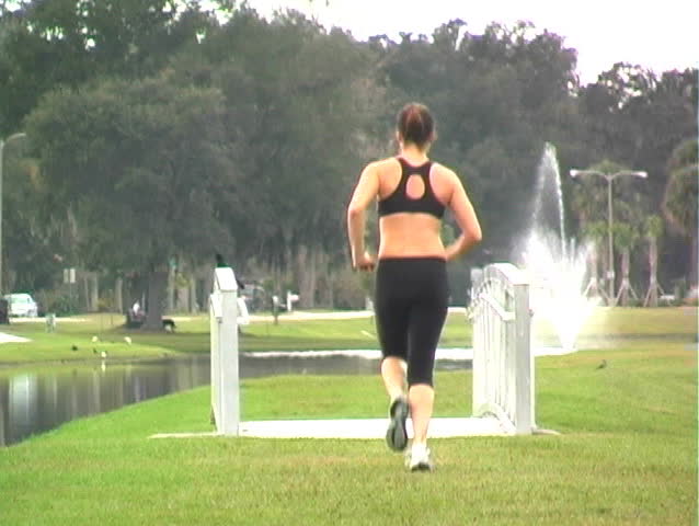 A lovely young brunette jogs away from the camera, then slows to a walk, across a foot bridge in a scenic municipal park with traffic and a water fountain in the background.