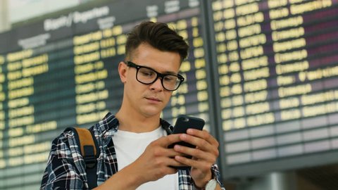 Portrait of young smiling man with glasses using smart phone for checking his flight on the arrivals table background.