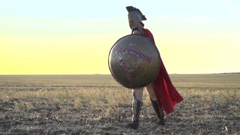 The majestic Roman legionary with a shield in his hand is standing in the field while in the wind his red cloak is blowing, slow motion