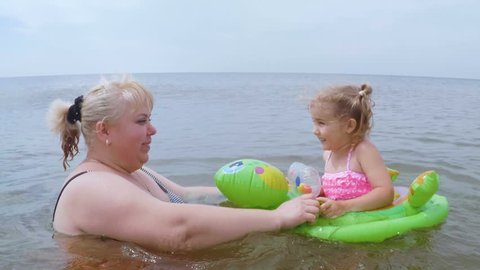  The mother and her little daughter bathe and play in the sea in the water. Mother with  kid swim in wavy sea, holding green inflatable ring sunny day.