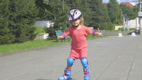 A little girl in a helmet and defense skates on roller skates. The child rolls on the rollers in the park.  Rollerblading In The Park.  Girl learns to ride a roller skate.