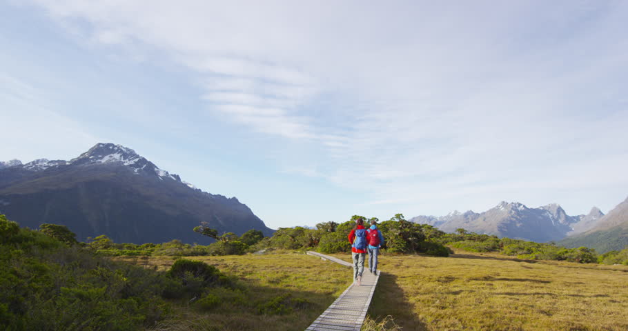 Male and female hikers walking on boardwalk at Routeburn rack. Couple on adventure is wearing backpacks while tramping on Key Summit Track during vacation at Fiordland National Park, New Zealand.