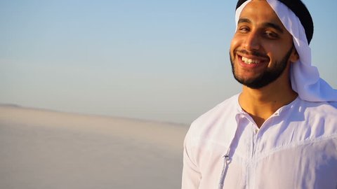 Close-up shot of portrait of Arabian Sheikh young man with beautiful smile that looks at camera and poses, standing among bottomless desert against blue sky on hot summer day in open air. Swarthy