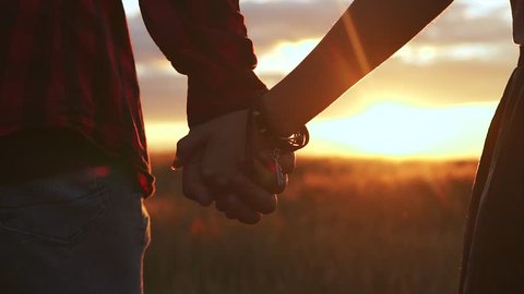 Close-up of hands joining together with sunset sunlight flare in the background. Beautiful romantic moment between two lovers walk in field. Concept of love of two people, relationship and lifestyle