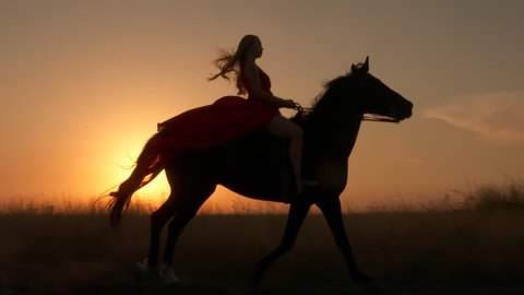 Young girl in red dress riding black horse against the sun. Rider with her stallion trotting across a field at sunset. Long gown blowing in the wind. Horseback riding in slow motion.